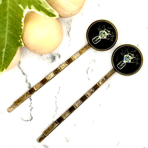 All Up In The Hair | Online Accessory Boutique Located in Mooresville, NC | Two Wildflower Bouquet Bobby Pins laying diagonally on a white marble background alongside a wood bead garland and ivy leaves.
