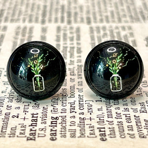 All Up In The Hair | Online Accessory Boutique Located in Mooresville, NC | Close up of two black Wildflower Earrings. The earrings are black with the image of a jar holding wildflowers and celestial details like a moon and saturn. The earrings are laying on a book page.