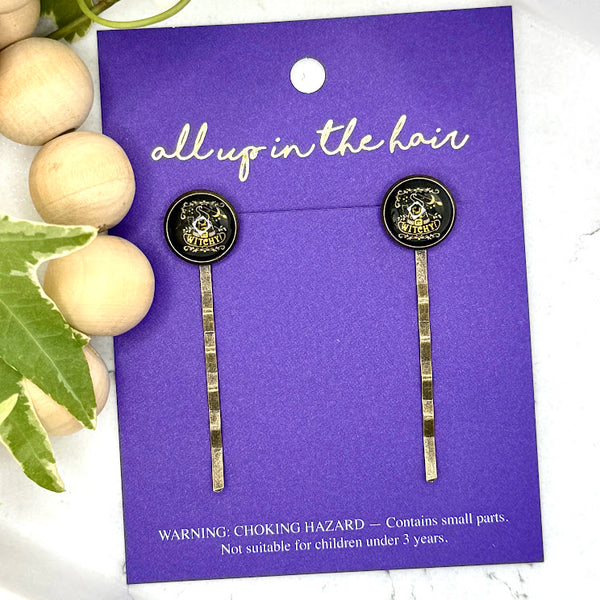 All Up In The Hair | Online Accessory Boutique Located in Mooresville, NC | Two Witchy Bobby Pins on an indigo colored, All Up In The Hair branded packaging card. The card is laying on a white marble background alongside a wood bead garland and ivy leaves.