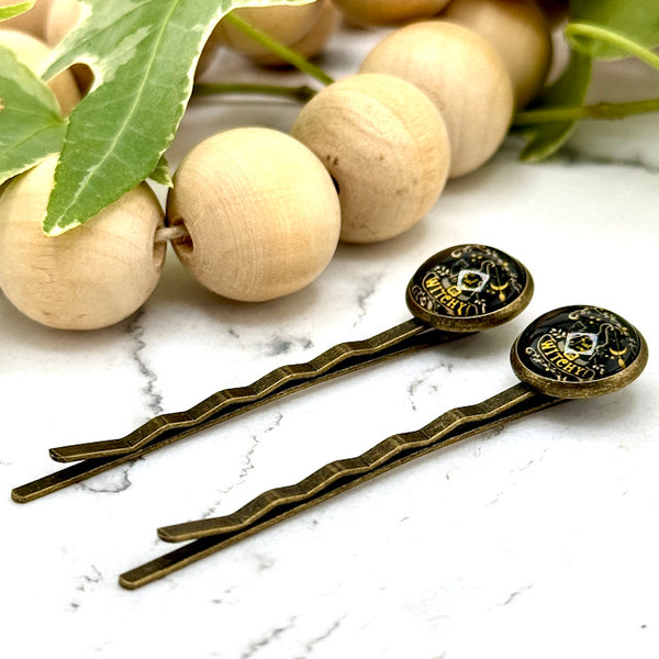 All Up In The Hair | Online Accessory Boutique Located in Mooresville, NC | Side view of two Witchy Bobby Pins on a white marble background alongside a wood bead garland and ivy leaves.