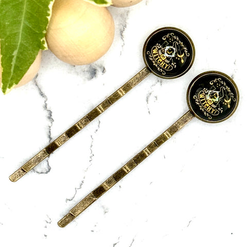 All Up In The Hair | Online Accessory Boutique Located in Mooresville, NC | Two Witchy Bobby Pins laying diagonally on a white marble background alongside a wood bead garland and ivy leaves.