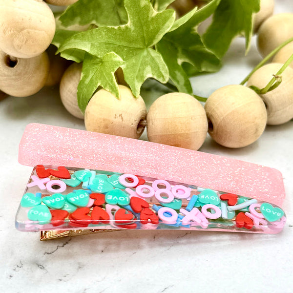 All Up In The Hair | Online Accessory Boutique Located in Mooresville, NC | Side view of our X's and O's Barrette Set laying on a white marble background. At the top of the image is a wood bead garland and ivy leaves.