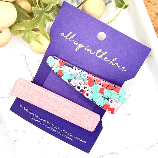 All Up In The Hair | Online Accessory Boutique Located in Mooresville, NC | Our X's and O's Barrette Set on indigo colored, All Up In The Hair branded packaging card. The card is laying on a white marble background. At the top of the image is a wood bead garland and ivy leaves.