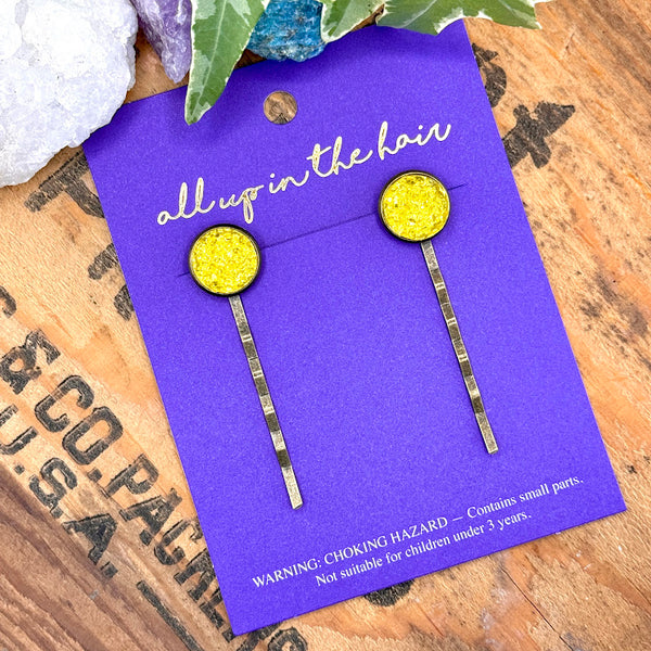 All Up In The Hair | Online Accessory Boutique Located in Mooresville, NC | Two Yellow Druzy Bobby Pins on an indigo colored, All Up In The Hair branded packaging card. The card is laying on a wood background with black lettering. There are crystals and ivy leaves at the top of the image.