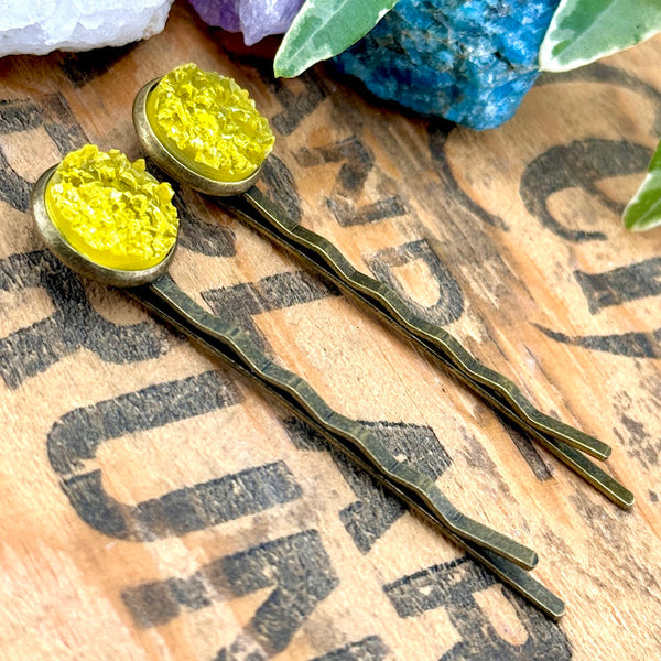 All Up In The Hair | Online Accessory Boutique Located in Mooresville, NC | Side view of two yellow druzy bobby pins laying on a wood background with black lettering. There are crystals and ivy leaves at the top of the image.