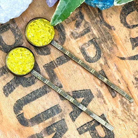 All Up In The Hair | Online Accessory Boutique Located in Mooresville, NC | Two Yellow Druzy Bobby Pins laying diagonally on a wood background with black lettering. There are crystals and ivy leaves at the top of the image.