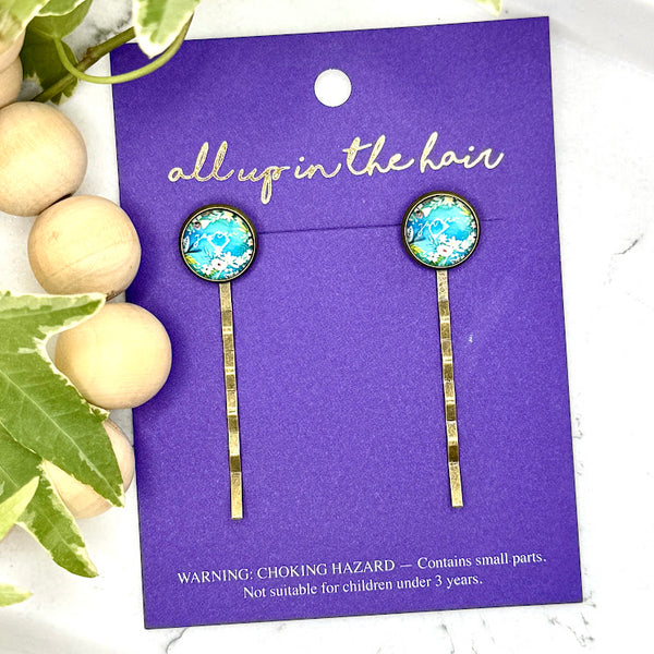 All Up In The Hair | Online Accessory Boutique Located in Mooresville, NC | Two You Can Bobby Pins on an indigo colored, All Up In The Hair branded packaging card. The card is laying on a white marble background alongside a wood bead garland and ivy leaves.