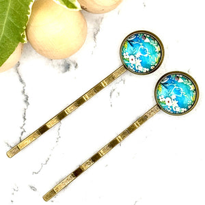 All Up In The Hair | Online Accessory Boutique Located in Mooresville, NC | Two Blue Floral Bobby Pins that say You Can laying on a white marble background alongside a wood bead garland and ivy leaves.
