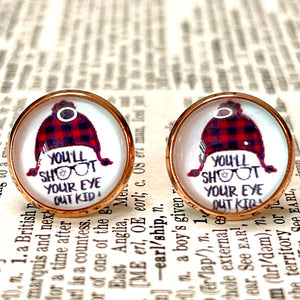 All Up In The Hair | Online Accessory Boutique Located in Mooresville, NC | Close up of a pair of our "You'll shoot your eye out" Earrings. The earrings feature a red buffalo plaid hat and the words " you'll shoot your eye out kid.' The earrings are laying on a book page.