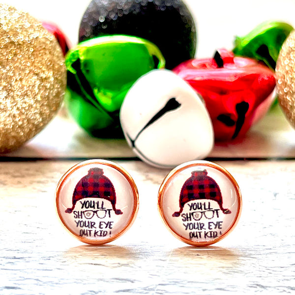 All Up In The Hair | Online Accessory Boutique Located in Mooresville, NC | Close up of a pair of our "You'll shoot your eye out" Earrings. The earrings feature a red buffalo plaid hat and the words " you'll shoot your eye out kid.' Behind the earrings are red, white, and green, jingle bells, along with black and gold glitter ornaments.