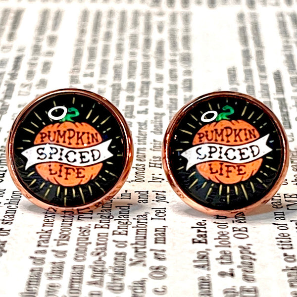 All Up In The Hair | Online Accessory Boutique Located in Mooresville, NC | Close up of two black earrings with a pumpkin in the center and the words "Pumpkin Spice Life". The earrings are laying on a book page.
